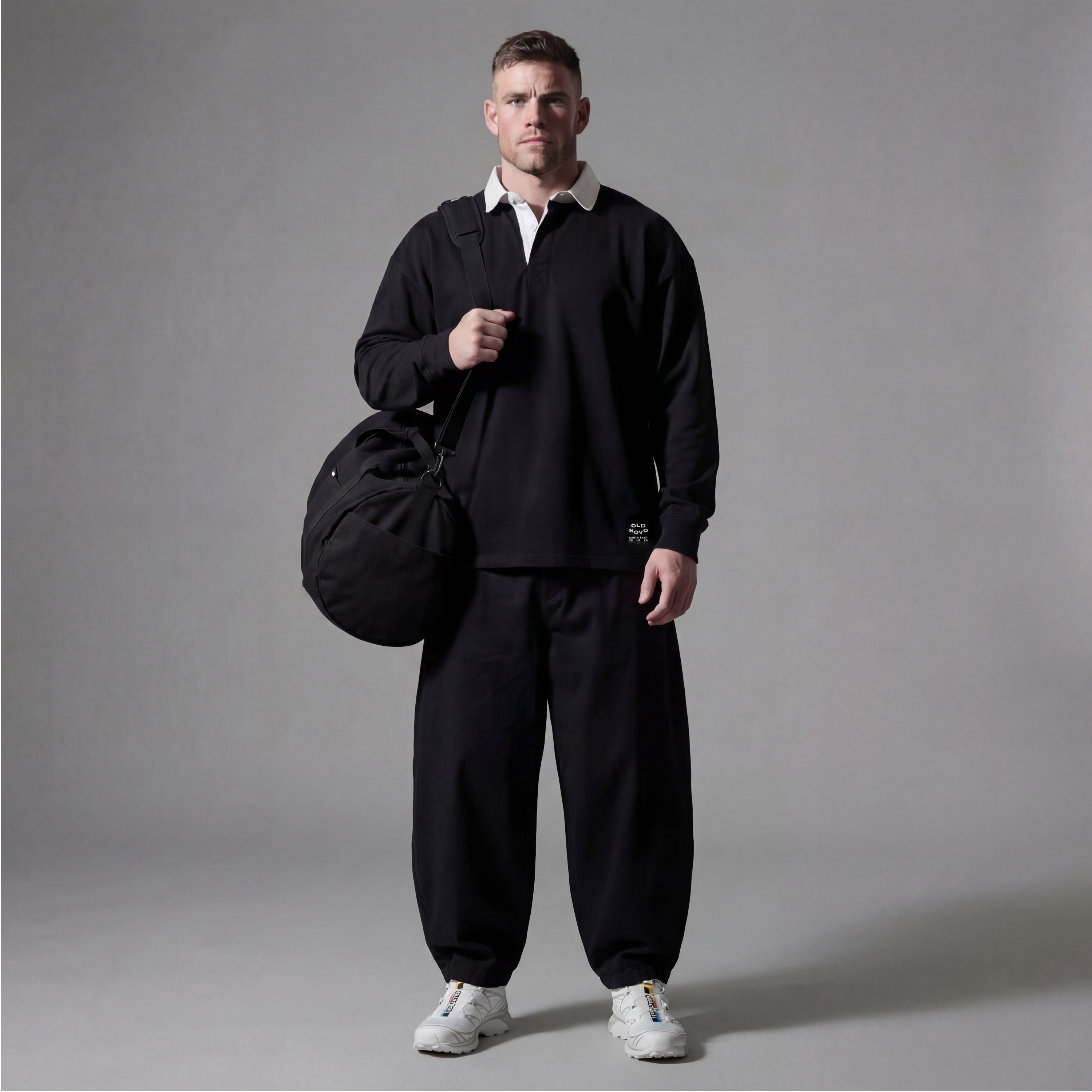 Man wearing a black tracksuit holding a black bag against a gray background